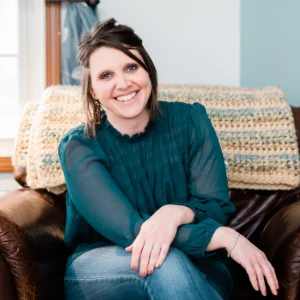 Alyssa Berthiaume, wearing a teal blouse and jeans, sitting on a brown chair with a crocheted blanket in the background.
