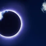 Solar eclipse with the moon covering the sun, surrounded by a glowing corona, partially obscured by clouds.