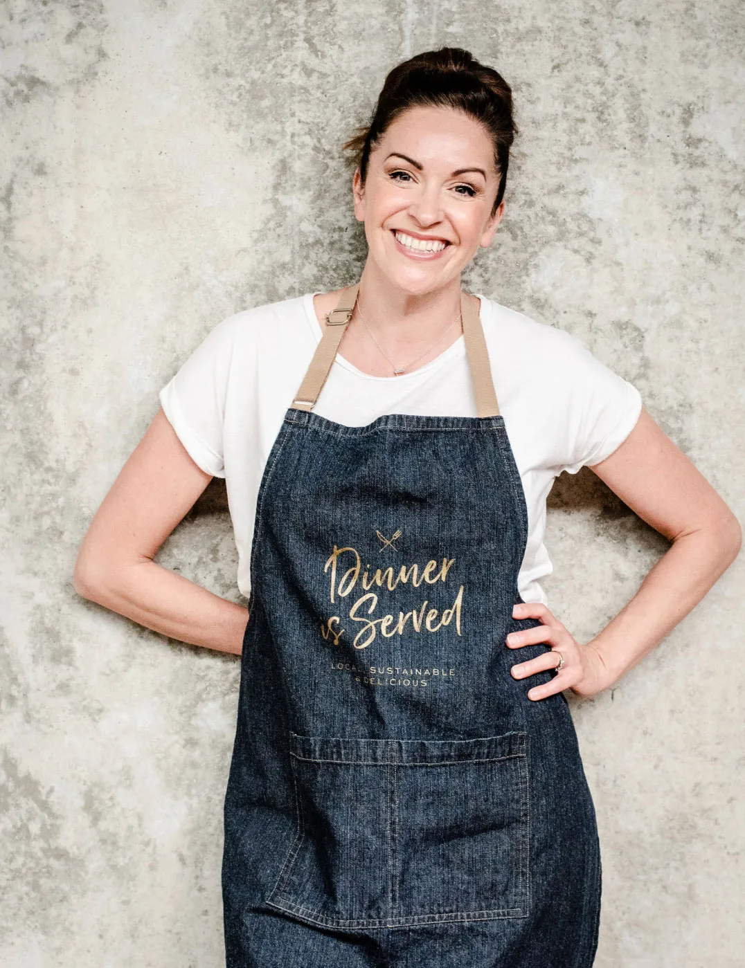 Claudia Lawrence, smiling and wearing a denim apron with 'Dinner is Served' printed on it, standing against a textured background.