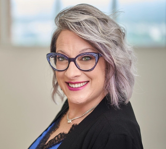Katie Nelson with silver-purple hair and statement glasses, smiling for a client testimonial about working with Kim Woods.