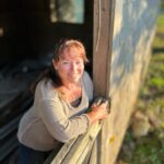start here Terrie Hanscom leans on a rustic wooden fence, smiling warmly in the sunlight, with a weathered wooden building and greenery in the background.