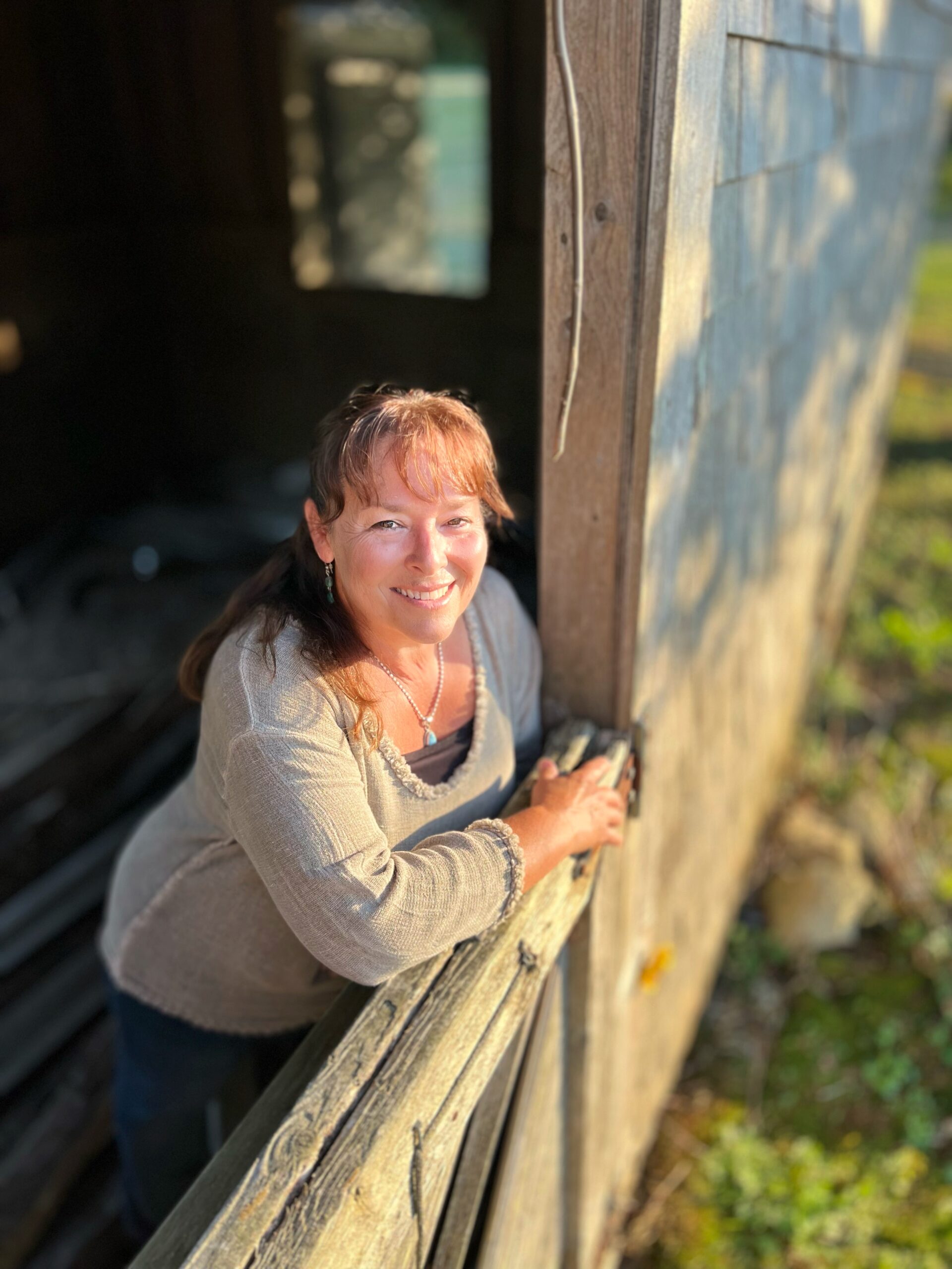Terrie Hanscom leans on a rustic wooden fence, smiling warmly in the sunlight, with a weathered wooden building and greenery in the background.