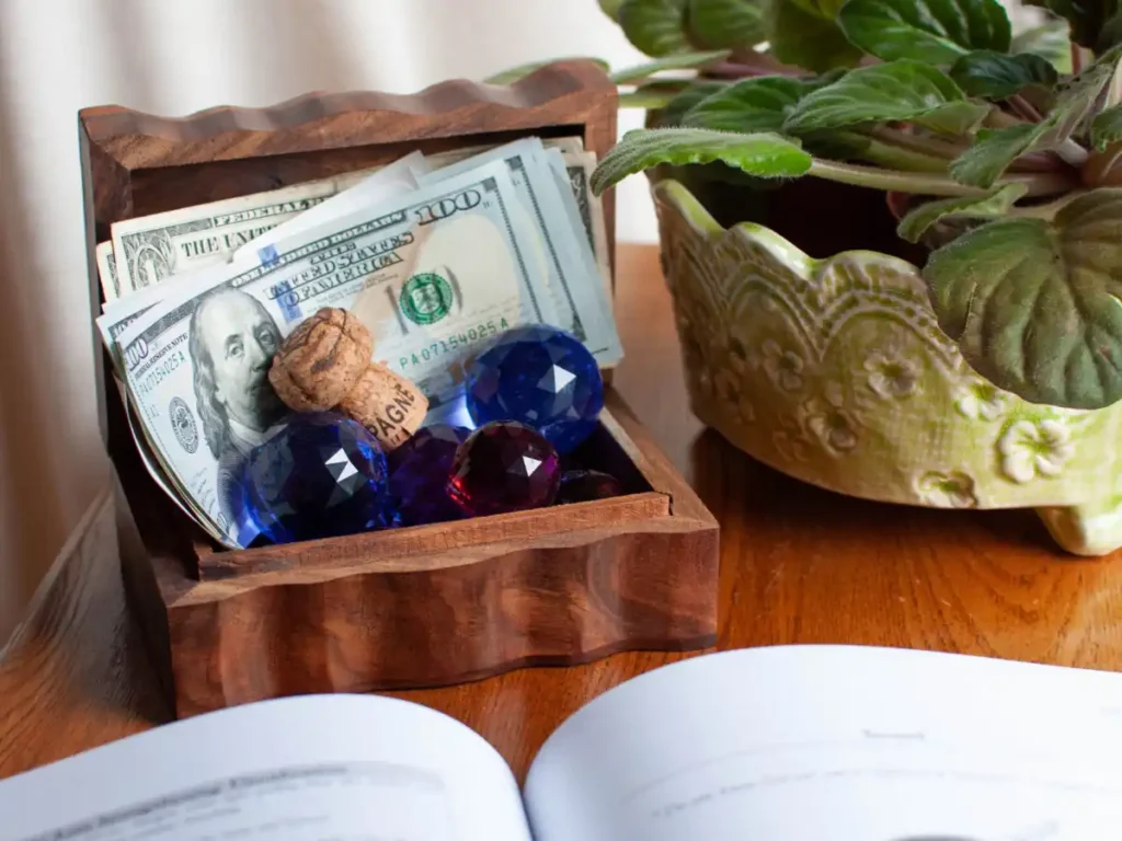 A small wooden box filled with hundred-dollar bills, colorful gemstones, and a champagne cork sits on a wooden table next to a green potted plant.