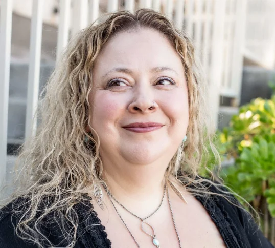 Crista G home page Smiling woman with curly, shoulder-length blonde hair wearing a black top and layered necklaces, standing outdoors in front of a light-colored railing and greenery.
