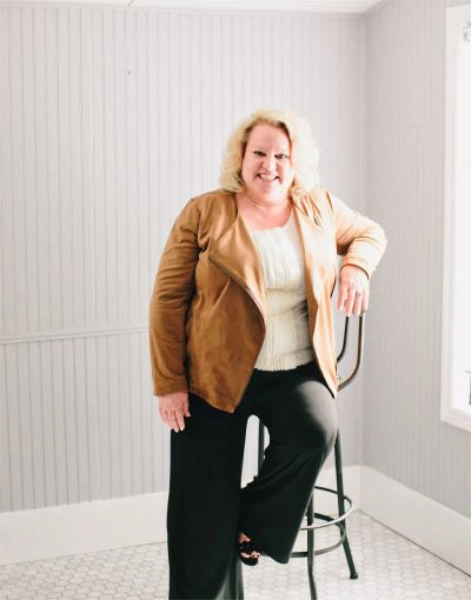 Kim Beige Standing Chair Kim Woods leaning casually on a tall chair, wearing a tan suede jacket, white blouse, and black pants, in a bright room with white paneled walls and natural light streaming through a window.