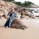 Kim Woods in a striped long cardigan and blue outfit sits barefoot on a rock at the beach, gazing into the distance. Waves crash against the rocky shoreline with cliffs and houses in the background.