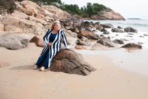 shop Kim Woods in a striped long cardigan and blue outfit sits barefoot on a rock at the beach, gazing into the distance. Waves crash against the rocky shoreline with cliffs and houses in the background.