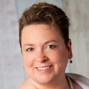 Testimonials Kristina Jensen with short hair and pearl jewelry in a softly lit, neutral-toned setting.