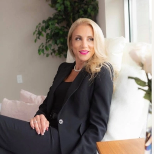Blonde woman in black suit smiling confidently while seated in a bright, modern space.