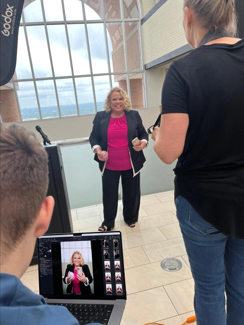 Kim Woods Business Strategist Master Astrologer Tower Hill PhotoShoot Behind-the-scenes photo of Kim Woods during a photoshoot, smiling in a bright pink top and black blazer, while a photographer captures her and a laptop screen shows her portraits in progress. Large arched windows provide natural light in the background.