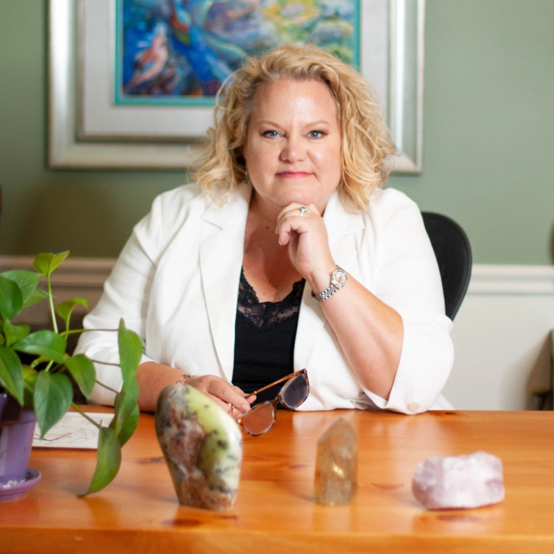 Kim Woods Business Strategist Master Astrologer sitting at her desk Kim Woods seated at a wooden desk with crystals and a potted plant, wearing a white blazer and looking confidently at the camera with her hand resting near her chin.