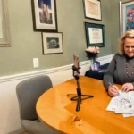 Kim Woods wearing a green sweater and pearl necklace writes on an astrological chart, with gemstones placed on the table. A smartphone on a tripod captures the scene, with framed certificates and artwork in the background.