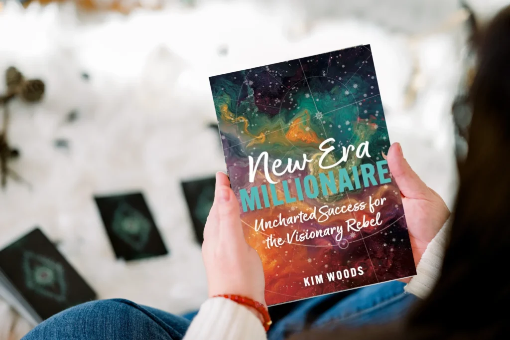 Person holding the book New Era Millionaire: Uncharted Success for the Visionary Rebel by Kim Woods, with tarot cards laid out on a white surface in the background.