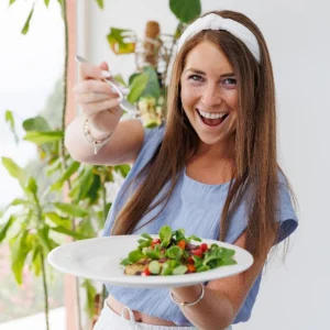Kim Woods Master Astrologer Client testimonial Hannah McKittrick Hannah McKittrick smiling and holding a plate of fresh salad, wearing a light blue top and white headband, surrounded by greenery.