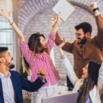 A diverse group of five cheerful professionals celebrating together in an office setting, raising their arms and smiling with excitement and joy.