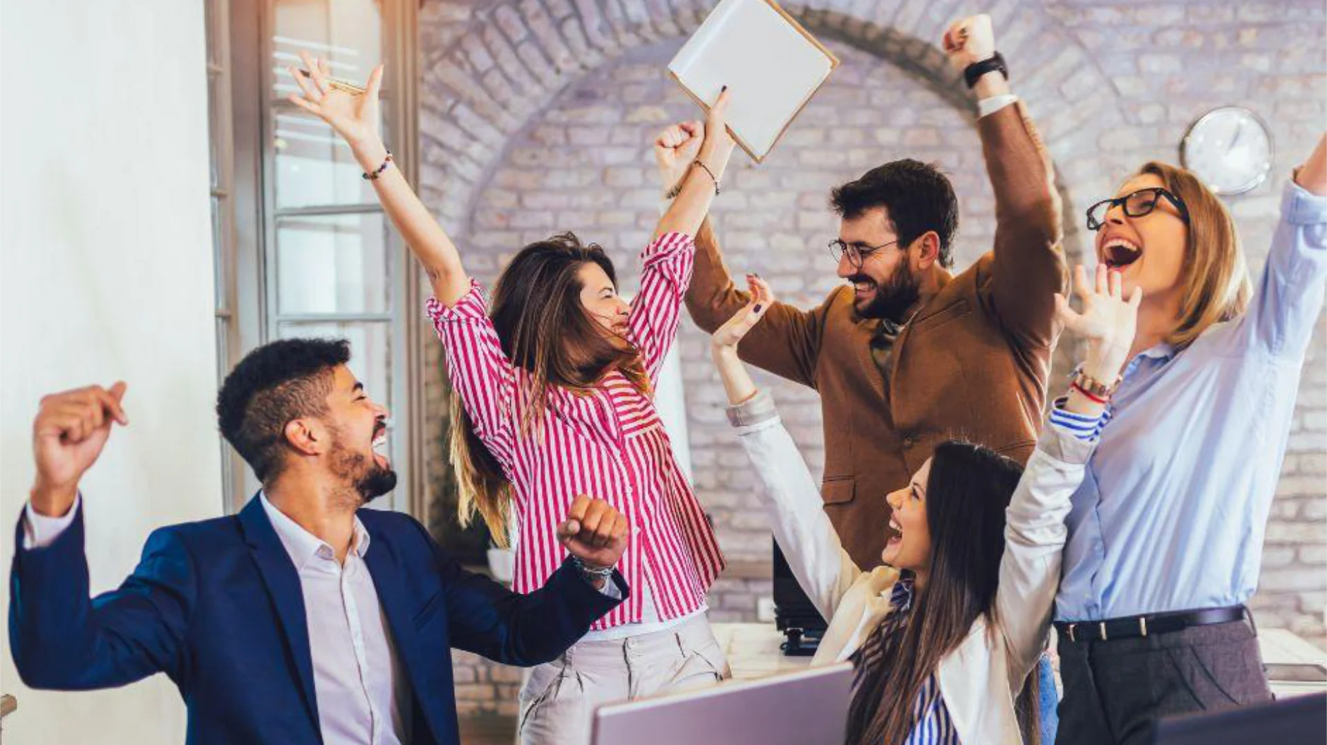 A diverse group of five cheerful professionals celebrating together in an office setting, raising their arms and smiling with excitement and joy.