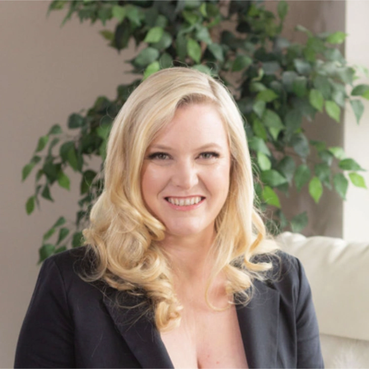 Hillary Gadsby Hillary Gadsby, smiling with long, wavy blonde hair, wearing a black blazer, seated indoors in front of a leafy green plant.