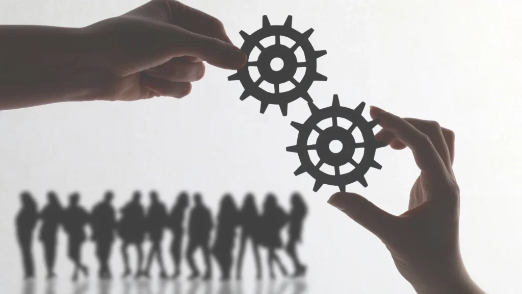 Two hands each holding a gear, with silhouetted people walking in the background, symbolizing alignment, teamwork, or systems in motion.