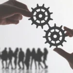 Two hands each holding a gear, with silhouetted people walking in the background, symbolizing alignment, teamwork, or systems in motion.