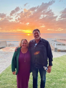 Kim Woods and Dan Vega smile together at sunset by the ocean, with warm colors in the sky and relaxed vibes.