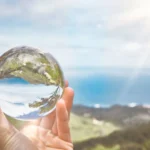 Hand holding a clear glass sphere reflecting a coastal landscape with ocean, hills, and sky in the background.