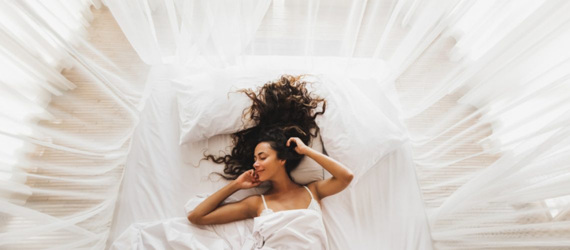 Beautiful smiling girl awakening in white bed. Happy wake up and start new day. Leisure and rest. View from above. Wellbeing concept.