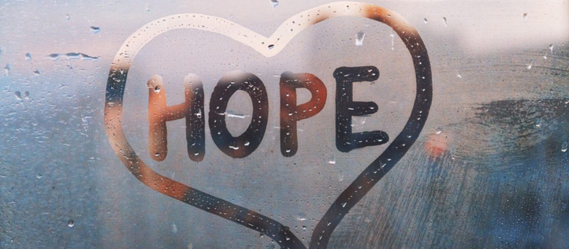 Handwritten word Hope in shape heart on misted glass on window flooded with raindrops on blue cloudy background